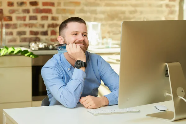 A happy man with a beard waits in front of the computer and holds a credit card at home. A guy doing an online payment on the internet on a desktop computer in his apartment.