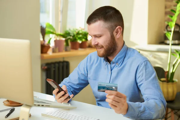 Sakallı ve sakallı mutlu bir adam akıllı telefondan ürünleri arıyor ve elinde evde bir kredi kartı tutuyor. Dairesindeki bir cep telefonuna internetten ödeme yapan bir adam..