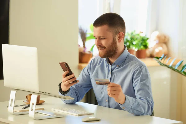 一个男人在他的智能手机上用信用卡在网上商店里找商品买东西 一个留着胡子的家伙在他公寓的笔记本电脑上上网付款 — 图库照片