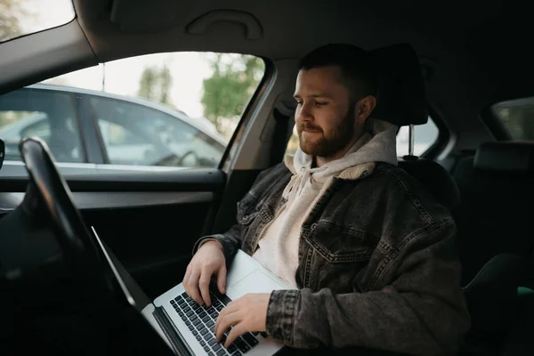 A happy man with a beard doing business online on his laptop computer inside a comfort car. A guy stopped his car to immediately remotely solve tasks at work in social distance.