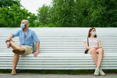 Kel bir adam ve genç bir kadın sıranın iki ucunda oturuyor. Koronavirüsün yayılmasını önlemek için birbirlerinden uzak duruyorlar. Bir adam ve yüzü maskeli bir kız parkta sosyal mesafede.