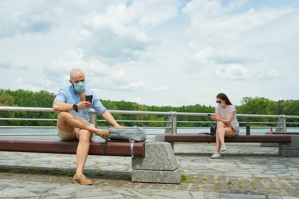 A bald man and a woman in medical face masks using smartphones keeping social distance sitting on different benches on a promenade. A guy and a girl resting in social distancing in the park.
