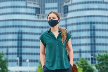 Tıbbi maske takmış bir kadın şehrin göbeğindeki pantolon ceplerine elini sokuyor. Sosyal mesafeyi koruyan kız, koronavirüsün yayılmasını önlemek için koruyucu yüz maskesi takıyor.