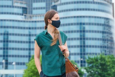 Tıbbi maske takan şık bir kadın elini pantolonunun cebine sokup şehir merkezinde bir çanta tutuyor. Sosyal mesafeyi koruyan kız, koronavirüsün yayılmasını önlemek için yüz maskesi takıyor.