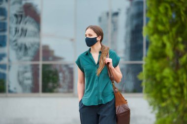 Tıbbi maske takan hoş bir kadın elini pantolonunun cebine sokup şehir merkezinde bir çanta tutuyor. Sosyal mesafeyi koruyan kız, koronavirüsün yayılmasını önlemek için koruyucu yüz maskesi takıyor.