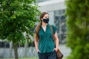 Tıbbi maske takan bir kadın ağaçların arasında yürüyor ve elleri şehir merkezinde pantolon ceplerine sokuyor. Sosyal mesafeyi koruyan bir kız, koronavirüsün yayılmasını önlemek için koruyucu yüz maskesi takar.