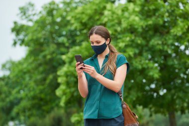 Lacivert tıbbi maske takan genç bir kadın parkta yürürken akıllı telefondan haber okuyor. Korunaklı maskeli bir kız ağaçların yakınındaki koronavirüsün yayılmasını önlemek için sosyal mesafeyi koruyor..