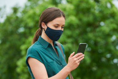 Tıbbi maske takan genç bir kadının yan görüntüsü parkta yürürken akıllı telefondan haber okuyor. Korunaklı maskeli bir kız, koronavirüsün yayılmasını önlemek için sosyal mesafeyi koruyor..