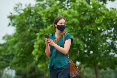 Parkta yürürken iki eliyle akıllı telefon tutan lacivert tıbbi maske takan genç bir kadın. Korunaklı maskeli bir kız, koronavirüsün yayılmasını önlemek için sosyal mesafeyi koruyor..