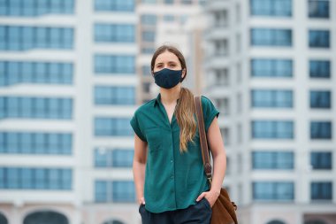 Tıbbi yüz maskesi takmış bir kadının portresi, şehrin ortasında yürürken ellerini pantolon ceplerine sokuyor. Sokakta sosyal mesafeyi koruyan koruyucu maskeli bir kız.