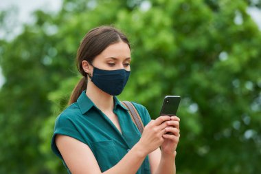 Tıbbi yüz maskesi takmış bir kadının portresi parkta yürürken akıllı telefondan haber okuyor. Korunaklı maskeli bir kız, koronavirüsün yayılmasını önlemek için sosyal mesafeyi koruyor..