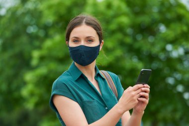 Tıbbi yüz maskesi takmış bir kadının portresi parkta yürürken bir akıllı telefon tutuyor. Korunaklı maskeli bir kız, koronavirüsün yayılmasını önlemek için sosyal mesafeyi koruyor..