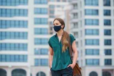 A pretty woman in a medical face mask is thrusting hands into pockets of trousers while walking in the center of the city. A girl in a protective mask keeping social distance on the street. clipart