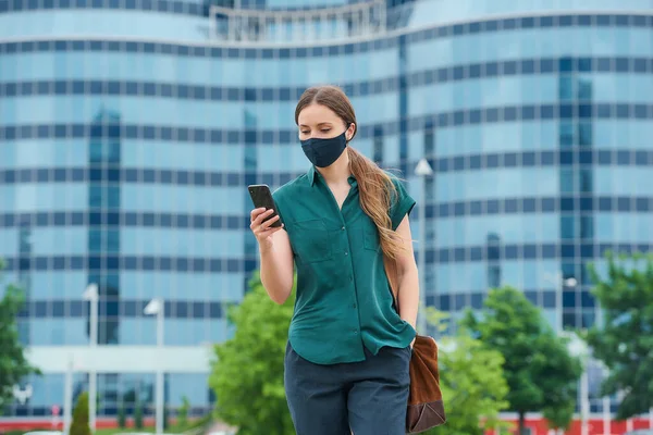 一名身穿海军蓝医疗面罩的年轻女子 以避免在市中心行走的头颈类动物的传播 一个长头发的女孩正在市中心用智能手机看新闻 — 图库照片