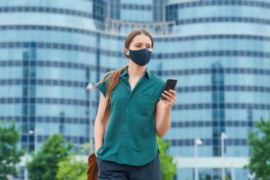 Şehrin merkezinde yürüyen koronavirüsün yayılmasını önlemek için tıbbi maske takan bir kadın. Uzun saçlı bir kız akıllı bir telefon tutuyor şehir merkezinde bir pantolon cebine elini sokuyor..
