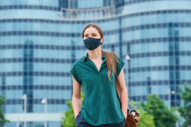A pretty young woman in a medical face mask to avoid the spread of coronavirus walking in the center of the city. A girl with long hair keeping social distance wears a protective face mask in downtown clipart