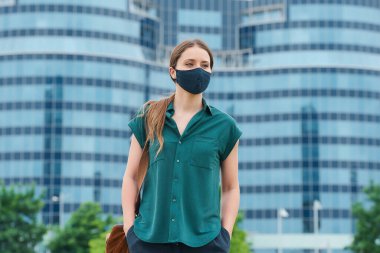 Tıbbi maske takmış bir kadın şehrin göbeğindeki pantolon ceplerine elini sokuyor. Sosyal mesafeyi koruyan kız, koronavirüsün yayılmasını önlemek için koruyucu yüz maskesi takıyor.