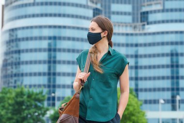 Tıbbi maske takan şık bir kadın elini pantolonunun cebine sokup şehir merkezinde bir çanta tutuyor. Sosyal mesafeyi koruyan kız, koronavirüsün yayılmasını önlemek için yüz maskesi takıyor.