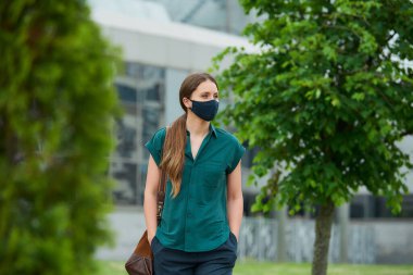 Tıbbi maskeli yalnız bir kadın ağaçların arasında yürüyor. Ağaçların arasından elini pantolonunun cebine sokuyor. Sosyal mesafeyi koruyan kız, koronavirüsün yayılmasını önlemek için koruyucu yüz maskesi takıyor.