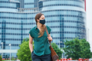 Tıbbi maskeli bir kadın elini pantolonunun cebine sokup şehrin ortasında deri bir çanta tutuyor. Kız sosyal mesafeyi koruyor. Koronavirüsün yayılmasını önlemek için koruyucu bir maske takıyor.