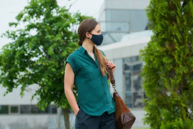 Tıbbi maske takan bir kadın ağaçların arasında yürüyor, elini pantolonun cebine sokuyor, deri bir çanta tutuyor ve şehir merkezine bakıyor. Sosyal mesafeyi koruyan koruyucu maskeli bir kız.