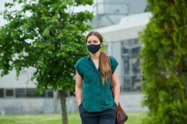 Tıbbi maske takan genç bir kadın ağaçların yanında yürüyor ve şehir merkezinde pantolon ceplerine elini sokuyor. Sosyal mesafeyi koruyan kız, koronavirüsün yayılmasını önlemek için koruyucu yüz maskesi takıyor.