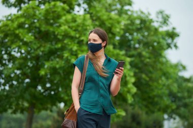 Lacivert tıbbi maske takan genç bir kadın, parkta yürürken pantolonunun cebine elini sokan akıllı bir telefondan haberleri okuyor. Koruyucu maskeli bir kız sosyal mesafeyi ağaçların yakınında tutuyor..