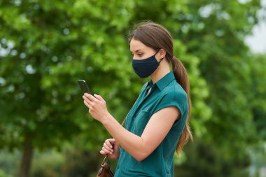 Tıbbi maske takan genç bir kadının yan görüntüsü, akıllı telefondan haberleri okumak ve parkta yürürken bir çanta tutmaktır. Ağaçlar arasındaki sosyal mesafeyi koruyan koruyucu maskeli bir kız..