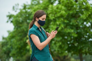 Tıbbi maske takmış bir kadının yan görüntüsü, parkta yürürken telefonla haber okuması ve bir çanta tutmasıdır. Korunaklı maskeli bir kız, koronavirüsün yayılmasını önlemek için sosyal mesafeyi koruyor.