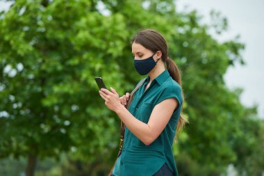 Tıbbi maske takmış bir kadının yan görüntüsü, parkta yürürken telefonla haber okuması ve bir çanta tutmasıdır. Korunaklı maskeli bir kız, koronavirüsün yayılmasını önlemek için sosyal mesafeyi koruyor.