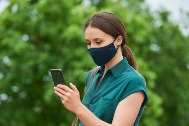 Tıbbi maske takan genç bir kadının yan görüntüsü parkta yürürken akıllı telefondan haber okuyor. Korunaklı maskeli bir kız, koronavirüsün yayılmasını önlemek için sosyal mesafeyi koruyor..