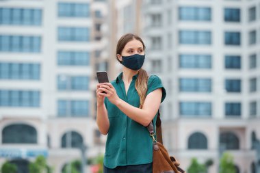 Tıbbi yüz maskesi takmış bir kadının portresi şehrin merkezinde yürürken bir akıllı telefon tutuyor. Korunaklı maskeli bir kız, koronavirüsün yayılmasını önlemek için sosyal mesafeyi koruyor..
