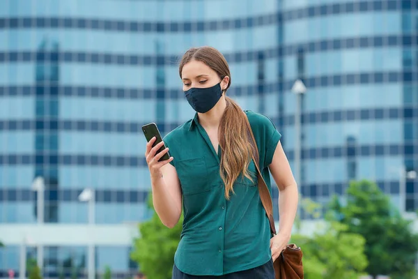 A woman in a medical face mask to avoid the spread of coronavirus in the center of the city. A girl with long hair reading the news on smartphone thrusting a hand into a pocket of trousers in downtown
