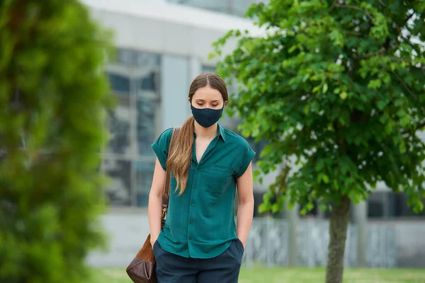 Sad woman in a medical face mask walks between trees thrusting hands into pockets of trousers in downtown. Girl keeping social distance wears a protective face mask to avoid the spread of coronavirus