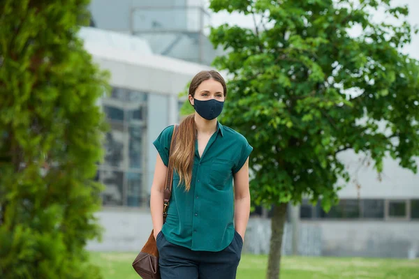 Tıbbi maske takan genç bir kadın ağaçların yanında yürüyor ve şehir merkezinde pantolon ceplerine elini sokuyor. Sosyal mesafeyi koruyan kız, koronavirüsün yayılmasını önlemek için koruyucu yüz maskesi takıyor.