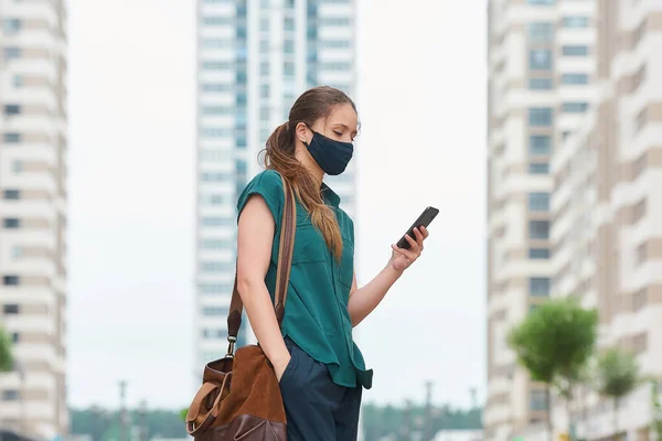 Side view of woman in a medical face mask who reads news on a smartphone thrusting a hand into a pocket of trousers walks between skyscrapers. Girl keeping social distance wearing protective face mask