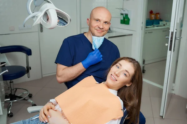 A bald dentist lowered the medical face mask near a smiling female patient in a dentist\'s office. A portrait of a doctor and a patient who demonstrated an excellent result of the tooth treatment.