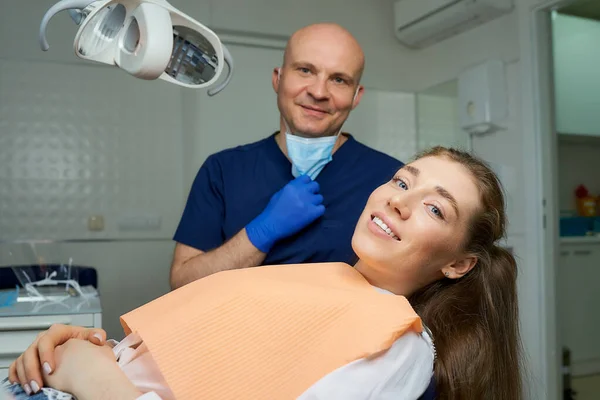 Dentist lowered the medical face mask near laying smiling female patient in dental chair. A doctor with a patient who is demonstrating an excellent result of the tooth treatment in a dentist\'s office