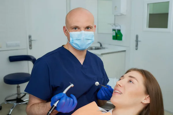 A dentist in a medical face mask holding a dental mirror and a drill before a procedure near a pretty girl in the dental chair. A doctor and a female patient during treatment in a dentist\'s office