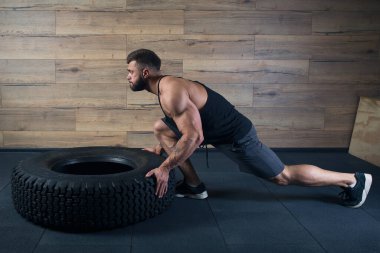 Dövmeli ve sakallı bir vücut geliştirici spor salonunda lastik itiyor. Siyah askılı ve gri şortlu kaslı bir adam çapraz atlet egzersizi yapıyor..
