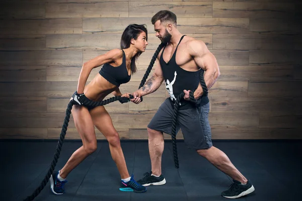 Sportif bir kız ve sakallı kaslı bir adam spor salonunda CrossFit eğitimi sırasında ip çekiyorlar. Dövmeli bir vücut geliştirici, siyah atlet ve şortlu bir fitness kızıyla antrenman yapıyor.