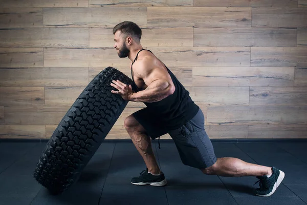 A strong man in a black tank top and grey shorts with a beard tries to push a tire in a gym. A muscular shredded guy doing a crossfit workout. A fellow with tattoos doing hardcore exercise.