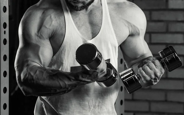 Close photo of arms of a bodybuilder who is doing biceps curls with dumbbells. A muscular man with tattoos and a beard in a white tank top and blue shorts is training in a gym. A black and white photo
