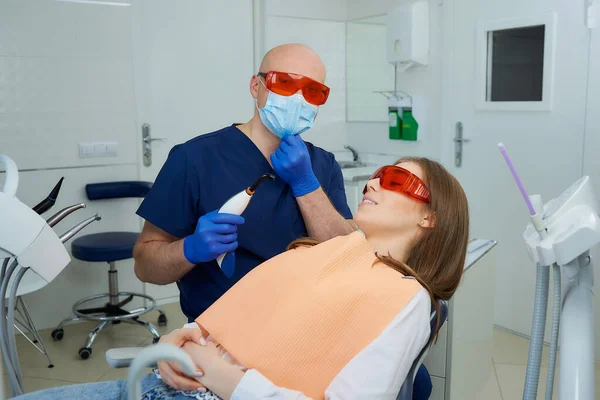 Dentist in UV protection glasses is checking a medical face mask and holding a dental polymerization light near girl in the dental chair. Doctor and female patient during treatment in dentist\'s office