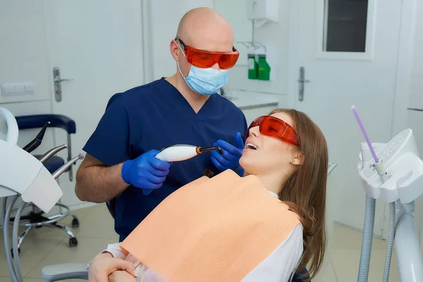 A dentist in a medical face mask and UV protection glasses holding a dental polymerization light near a woman in the dental chair. A doctor and a patient before treatment in a dentist\'s office.