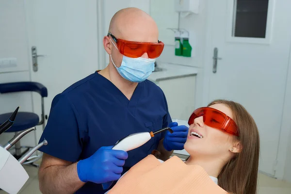 A dentist in a medical face mask and UV protection glasses holding a dental polymerization light near a young woman in the dental chair. A doctor and a patient started treatment in a dentist\'s office.