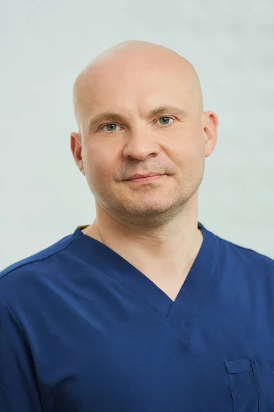 A close portrait of a caucasian bald serious adult dentist in the navy blue uniform is posing in a dentist\'s office.