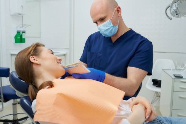 Orta yaşlı kel bir dişçi dişçi dişçi muayenehanesinde beyazlatma işleminden önce gülümseyen bir kadın hasta için diş gölgesi seçer. Bir doktor, tedavi öncesi diş rengini bir hastayla eşleştiriyor..