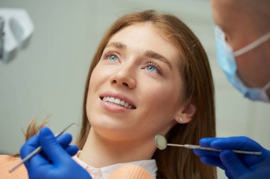 Gülümseyen güzel bir kadın, diş aynası ve dişçi koltuğunda bir diş kâşifi ile tıbbi yüz maskesi takan bir dişçi tarafından muayene ediliyor. Hastanın dişlerini dişçinin ofisinde tedavi eden bir doktor..