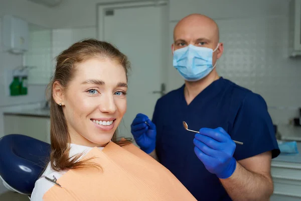 Dentist in medical face mask is holding a dental mirror and dental explorer near a laying female patient in a dental chair. A doctor with a patient who is demonstrating a result of the tooth treatment